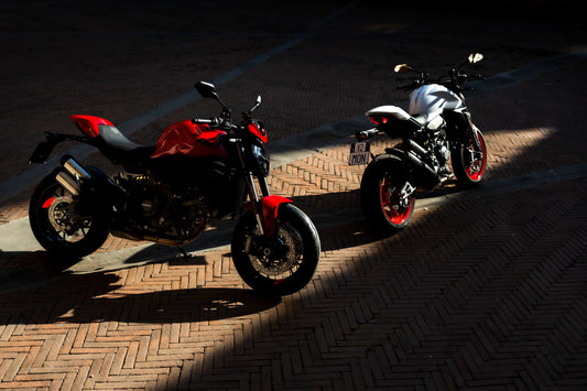 Two 2026 Ducati Monster's, one red and one white sitting on sun streaked pavement