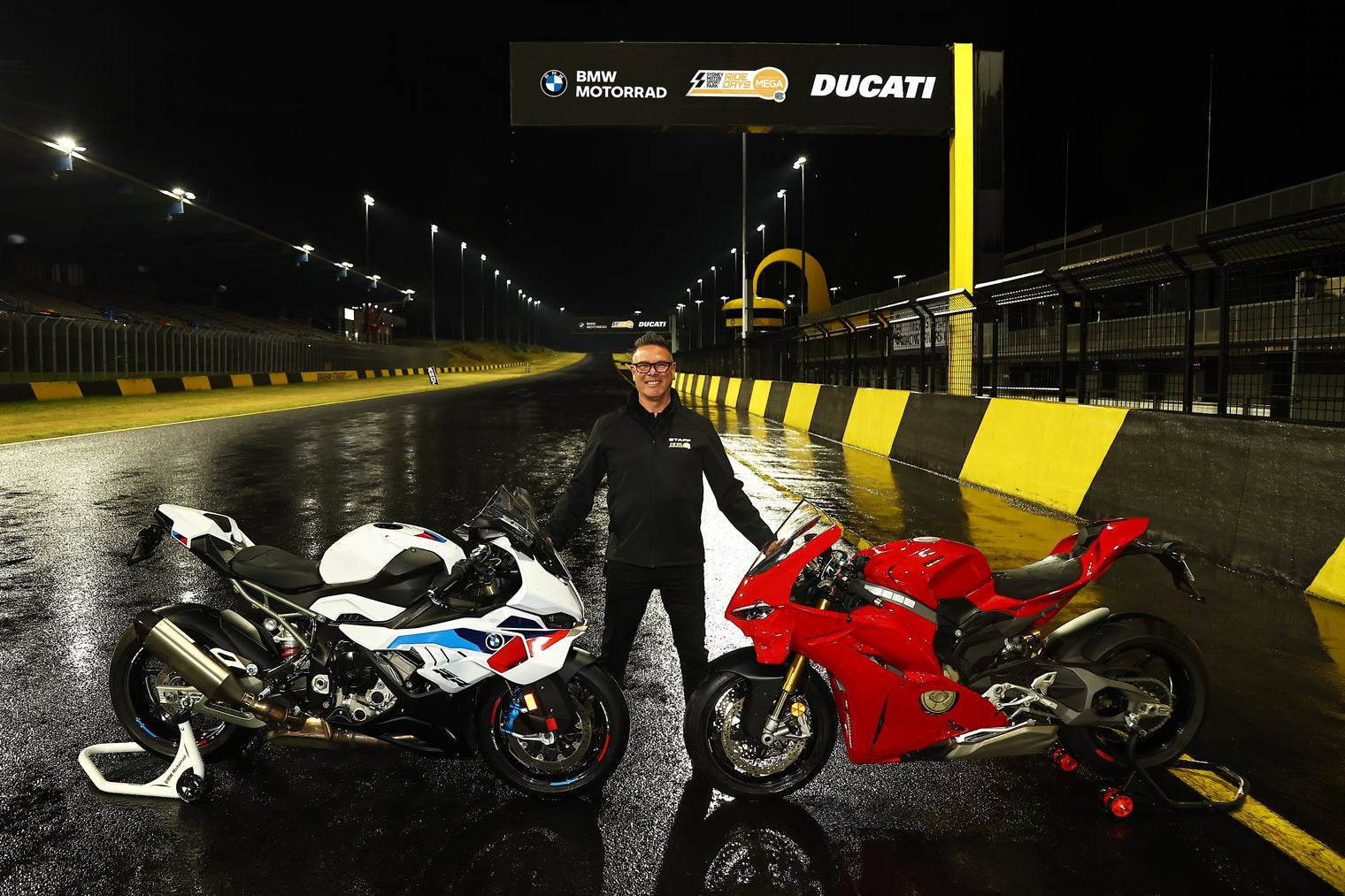Man with a BMW S1000RR and a Ducati Panigale V4S standing at night with the bikes on the straight at Sydney Motorsport Park.