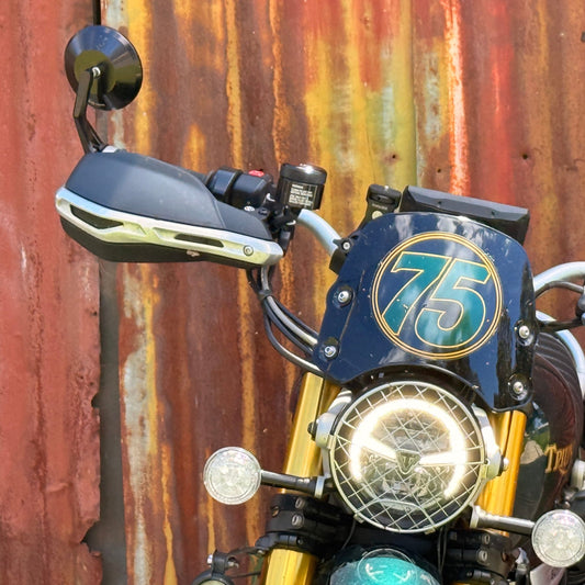 Close-up of a motorcycle front end with a blue windscreen featuring a gold '75' emblem, gold forks, round headlights, and dark handlebar covers, set against a rusted corrugated metal wall.